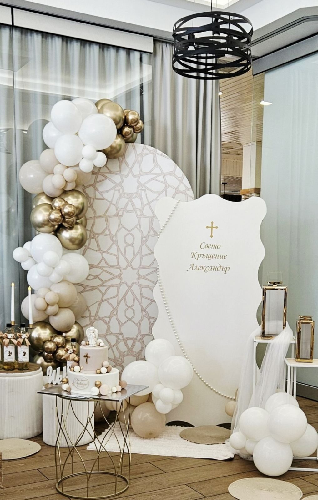 Elegant balloon decor in white, cream and gold chrome around a round backdrop with a geometric pattern, a pearl-trimmed sign, cake and lanterns